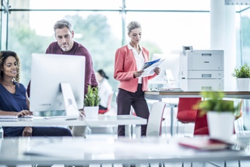 Woman looking at a document while stood next to a brother printer and man and woman looking at a pc screen